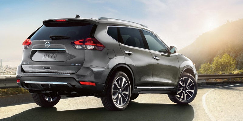 A gray Nissan Rogue SUV parked on a mountain road overlooking a city at sunset.