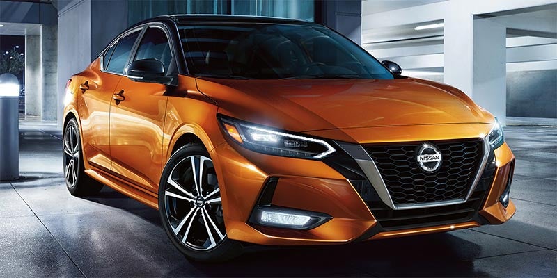 Front 3/4 view of an orange Nissan car with a black roof, black Nissan logo, and bright headlights.