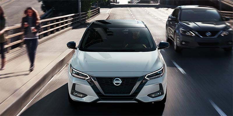 White parked Nissan Sentra, black roof, with two blurred people walking past, and a dark grey car driving by.