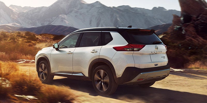 White Nissan Rogue SUV driving on a dirt road, viewed from the rear, with rocky mountains in the background