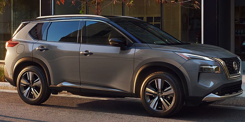 A side view of a gray Nissan Rogue parked on a paved street, in front of a building with bright windows.