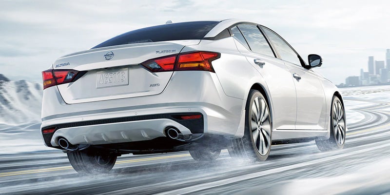 Rear view of a white Nissan Altima with AWD driving on a wet road, tire treads visible, with a city in the distance