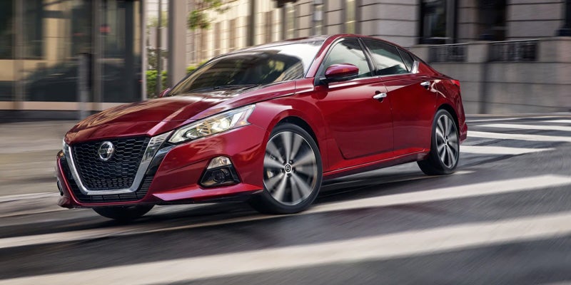 A shiny red Nissan Altima sedan drives right to left across a blurred street
