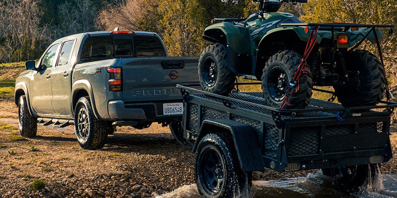 Gray pickup truck towing a black utility trailer with a green ATV on it, crossing a shallow creek in a wooded area.