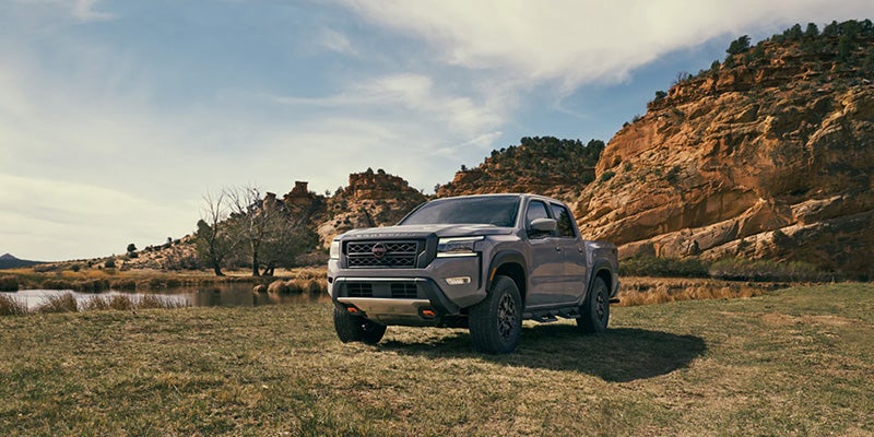 A dark gray Nissan Frontier Pro-4X truck on a grassy lakeside in a desert canyon under a partly cloudy sky
