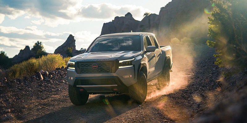 A grey Nissan Frontiers pickup truck drives on a dusty dirt road with large rocks and brush, mountains in background.