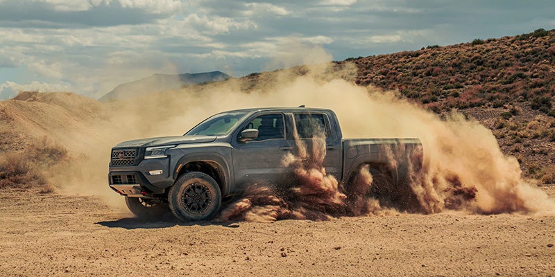 A dark gray off-road truck kicks up a large cloud of dust while driving left across a desert landscape.