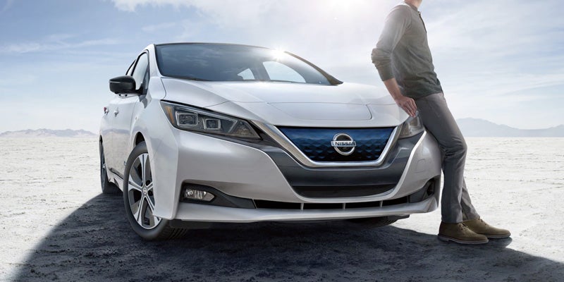 A man in gray casually leans against a white electric Nissan Leaf