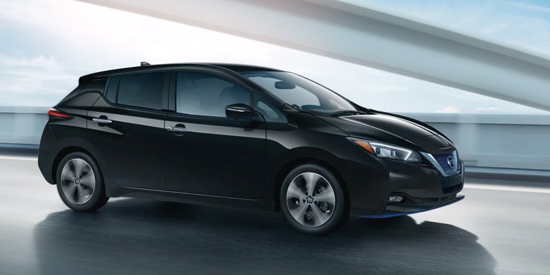 A black Nissan Leaf electric car driving on a multi-lane highway, with light-colored barriers and sky in the background