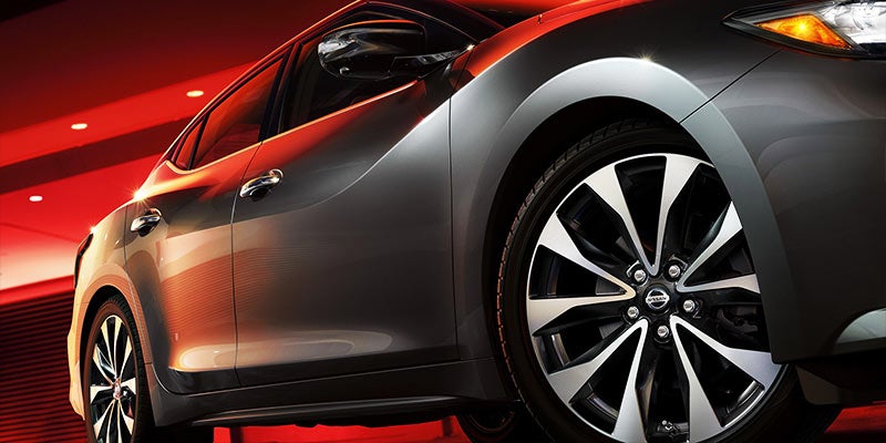 Side view of a dark gray Nissan sedan with black and silver multi-spoke wheels, bathed in a red light.