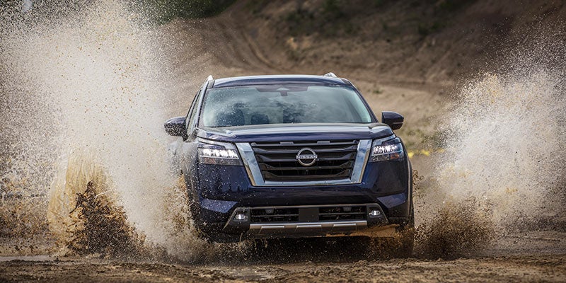 A dark blue SUV splashes through a mud puddle on a dirt road, spraying water and mud into the air