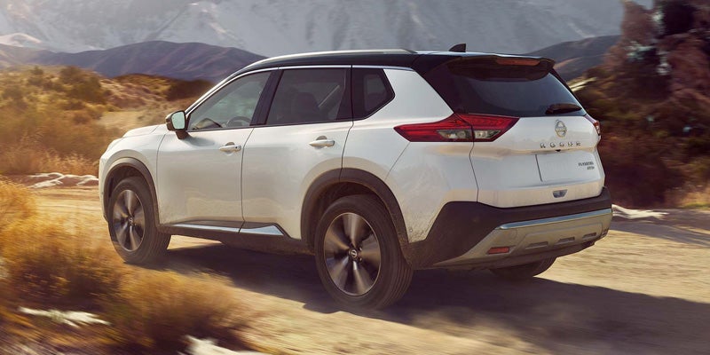 Rear view of a white Nissan Rogue with a black roof, driving on a dirt road through a desert landscape