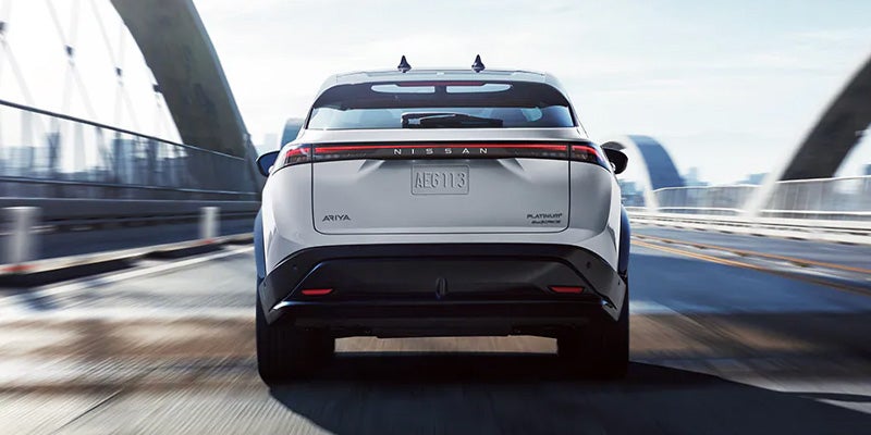Rear view of a white Nissan Ariya electric SUV driving on a bridge, with blurry city background
