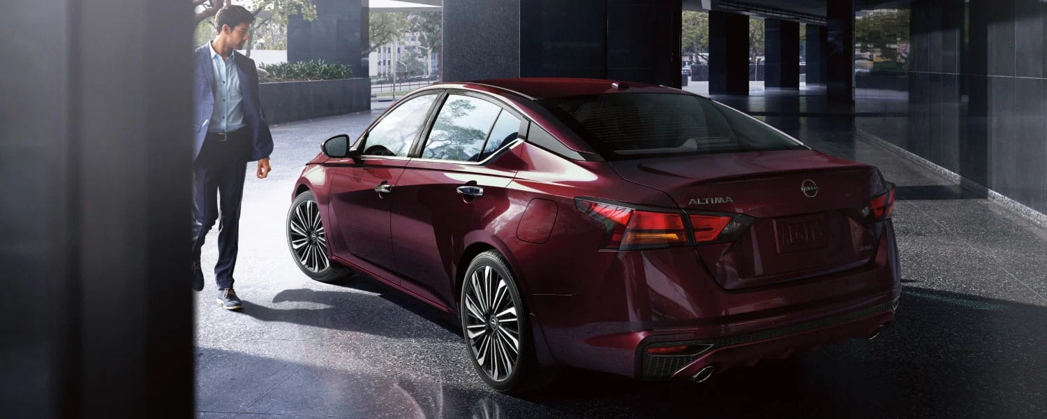 A man in a suit walks alongside a parked dark red Nissan Altima sedan
