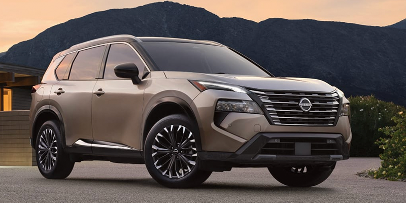 A gold Nissan Rogue parked in front of mountains with green bushes and a modern building under a sunset sky.