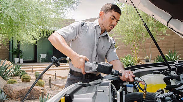 A technician in a light gray uniform charging a car battery under the hood of a white vehicle.