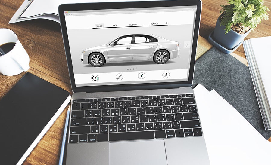 Laptop on a messy wooden desk, displaying a white car on an automotive services website.