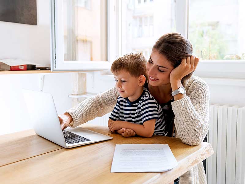A smiling woman with her young son using a laptop together at a wooden table in a bright room.